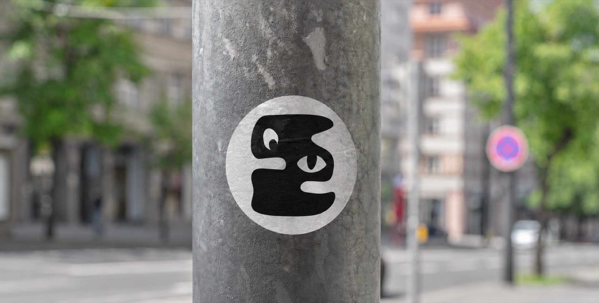 Der Sticker im Stadtraum: auf einen Laternenmast geklebt, dahinter unscharf eine Strasse mit Baeumen und Altbauten